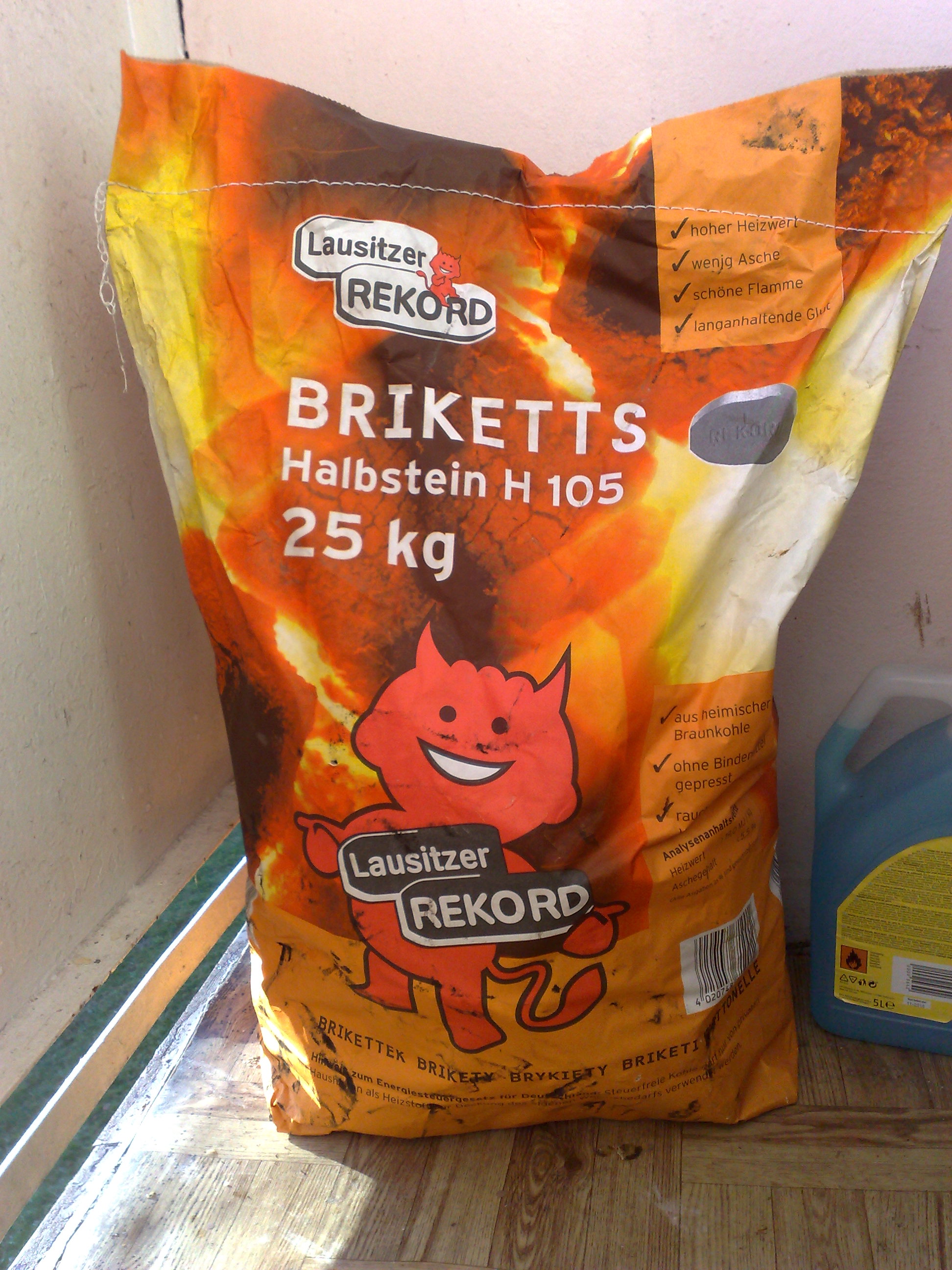 Żółto-pomarańczowy worek 25 kg z brykietami węgla brunatnego marki Lausitzer Rekord, z ilustracją czerwonego diabełka, stojący na podłodze, obok niebieskiego pojemnika z płynem.